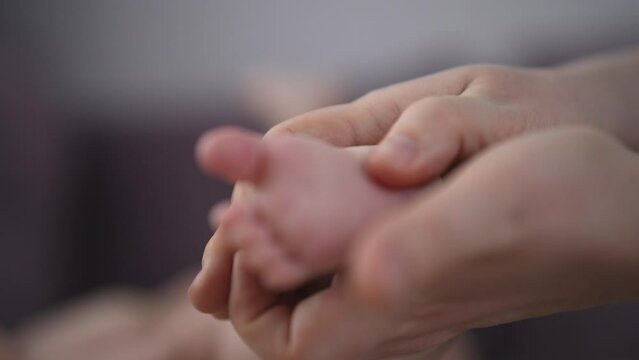 Close-up cropped shot of loving mother massaging newborn infant baby feet with thumbs tender touch parenting. Closeup of young mom touching legs of little infant kid lying on bed at home, slow motion.