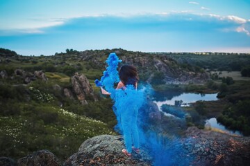 Beautiful young woman in Nature with Blue Smoke in the Air. The light and the shadow create a dramatic effect. The person is holding a blue smoke bomb.