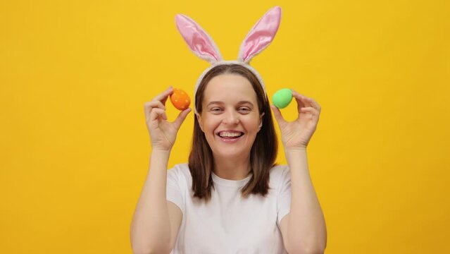 Joyful Easter holiday. Spring celebration traditions. Caucasian woman wearing rabbit ears headband holding colored easter eggs expressing happiness posing isolated over yellow background