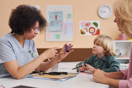 Side view portrait of caring Black woman as doctor showing inhaler to child in pediatricians office at clinic