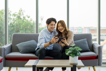 Young asian couple smiling and looking at a tablet on a grey sofa.