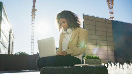 A young professional is immersed in her work on a laptop, seated outdoors with the sun casting a...