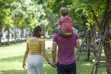 A man and woman are walking together holding hands in a beautiful green park, while the father holds a giggling small child on his shoulder. View from behind