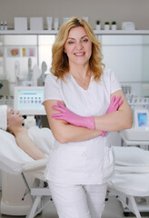 Professional Aesthetician in Clinic with Client in Background. Confident female aesthetician in white uniform and pink gloves stands with arms crossed in a modern beauty clinic with a client 