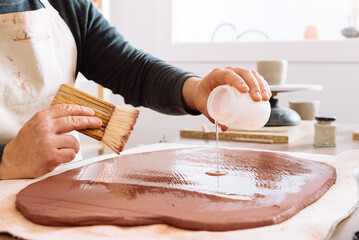 Crop artisan pouring liquid on clay slab
