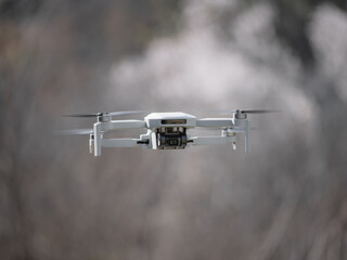 Small white drone with a built-in camera in mid-flight and a blurred background of trees with white flowers giving the sensation of smoke around and speed in general