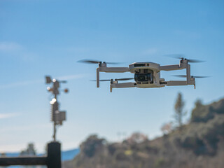 Small white drone with a built-in camera flies over a small weather station that shows the direction of the wind located on top of a wooden post and a blue sky in the background surrounded by mountain - obrazy, fototapety, plakaty