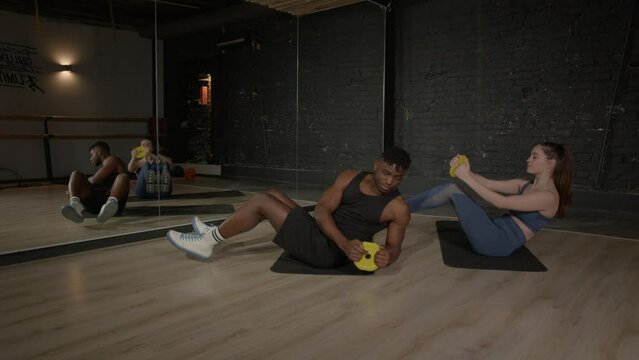 Active sporty muscular build black man and pretty female fitness trainer doing cross training workout together, practicing seated weight plate trunk rotation exercise in v-up position on yoga mats