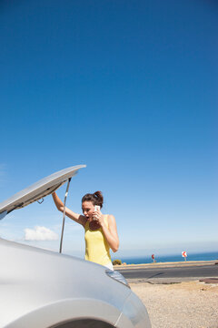Woman Looking under Car Hood whilst Talking on the Phone
