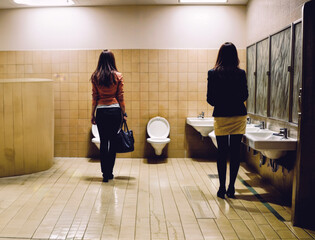 Two women in the women's restroom. The women stand with their backs to the viewer.