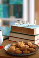 Cup of tea or coffee, pile of books, plate of cookies, reading glasses, e-reader and pen on the table. Colorful rainbow bookshelf in the background. Selective focus.