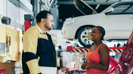 Asian automotive mechanic walk and talking to black woman customer about explain problem car condition to client in garage, auto service, maintenance and repair car engine, transportation concept. - obrazy, fototapety, plakaty