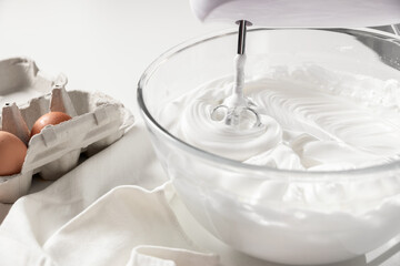 Close-up of a glass bowl filled with meringue and an egg beater on white table