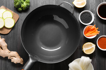 Empty iron wok surrounded by ingredients on dark grey wooden table, flat lay