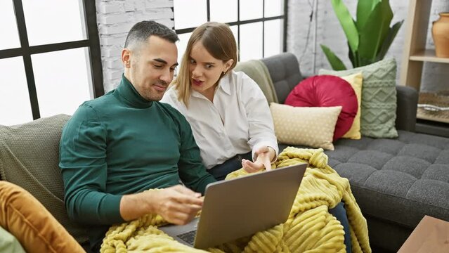 A woman and a man cuddle under a yellow blanket while happily using a laptop in a cozy, well-decorated living room.