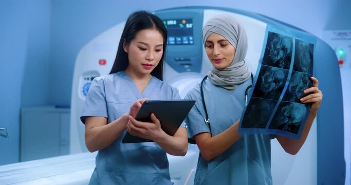 Doctor in hijab looking at MRI image. Muslim doctor consulting with intern. Nurse talking to chef doctor. Clinic of magnetic resonance tomography. Nurse holding folder and making notes.