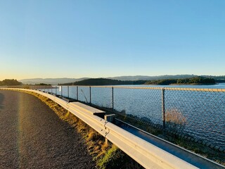 Fototapeta premium A road with a railing alongside a beautiful forest field and a serene waterscape during sunset