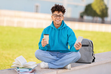 Cheerful student guy sitting with cellphone and takeaway coffee outdoor