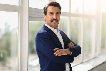 Fototapeta premium Confident middle-aged businessman in a blue suit with crossed arms smiling in a well-lit office