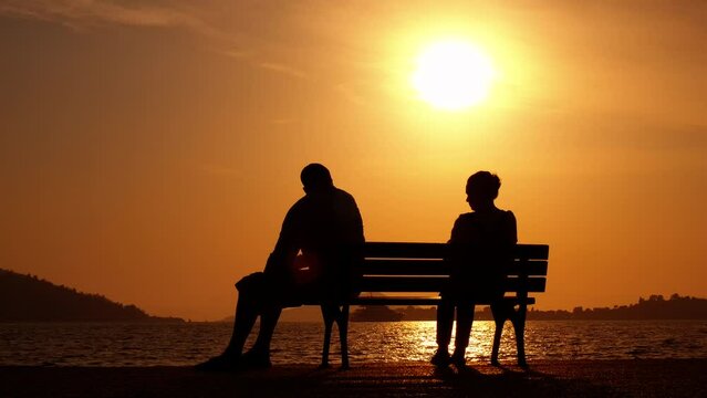 Quarrelled couple on bench in evening. A view of quarrelled couple silhouettes sitting on bench against evening lake water.