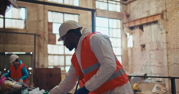 A man with Black skin in a special white protective uniform and an orange vest sorts plastic at an old waste processing plant. A man with Black skin is an employee of a huge waste recycling plant