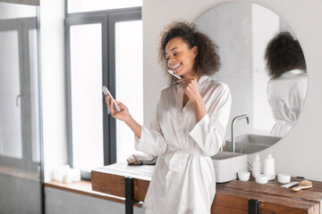 black lady getting ready brushing teeth using phone in bathroom - obrazy, fototapety, plakaty