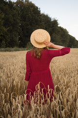 Back view portrait of young woman walking across wheap field. Lady wearing straw hat and boho style red dress.