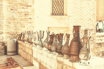 Row of aged clay jugs on a stone ledge against a brick wall, with a traditional rug on the floor and a patterned window in the background. Old photo style. Bukhara (Buxoro), Uzbekistan
