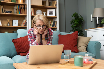 Worried senior woman sitting alone at home browsing bad news at internet. Old people reading online...
