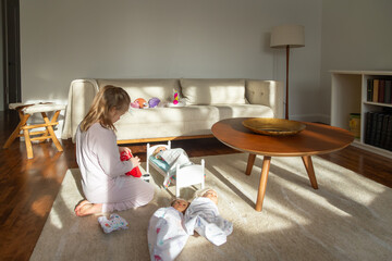 Five-year old little girl in pink pyjamas kneeling on carpet playing with her dolls in sun-filled...