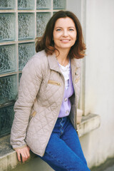 Outdoor portrait of happy 40 year old woman posing outside wearing beige jacket, leaning on window - 752174340