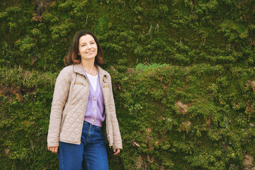 Outdoor portrait of happy beautiful woman enjoying nice day outside, posing with moss wall - 752174367