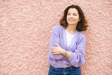 Outdoor portrait of pretty 40 - 45 year old woman leaning on pink wall, wearing purple cardigan, spring mood and colours - 752174399