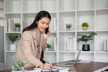 Young Asian business woman sits on the phone in an online business meeting using a laptop in a modern home office decorated with shady green plants.