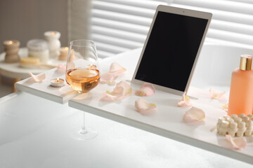 Wooden tray with tablet, wine, toiletries and flower petals on bathtub in bathroom