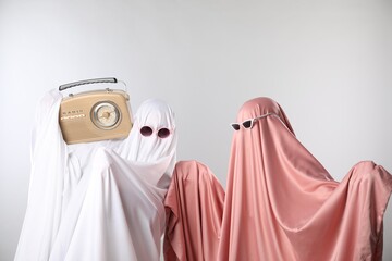 Glamorous ghosts. Women in color sheets and sunglasses with retro radio receiver on light grey background
