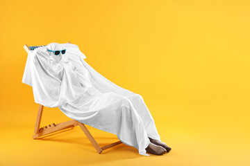 Person in ghost costume and sunglasses relaxing on chaise longue against yellow background, space...