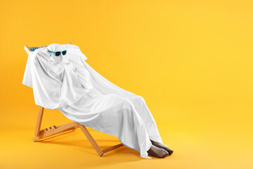 Person in ghost costume and sunglasses relaxing on chaise longue against yellow background, space for text
