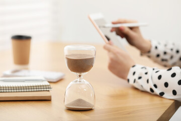 Hourglass with flowing sand on desk. Woman using tablet indoors, selective focus