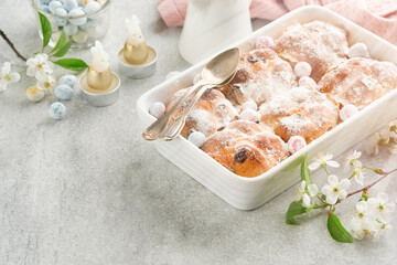 Easter Hot cross buns. Traditional Easter treats cross buns with raisins, butter, chocolate candy eggs with blossoming cherry or apple flowers on rustic light wooden backgrounds. Easter holiday meal.