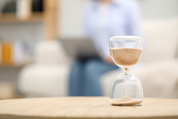 Hourglass with flowing sand on desk. Woman using laptop indoors, selective focus