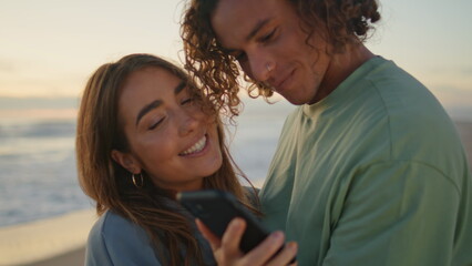 Love pair looking smartphone sunrise sea closeup. Young man kissing girlfriend