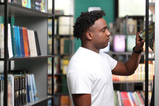 Portrait of young African American man taking book from shelf in university library to study for exam. Cute dark-skinned man in white t-shirt with hand picking interesting book from shelf, side view