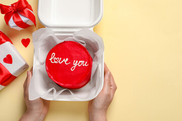 Woman holding takeaway box with bento cake at beige table, top view. St. Valentine's day surprise