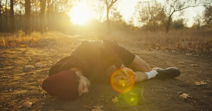 Sad tired guy athletes in a red cap in a black sports uniform falls to the ground while hurting his leg holds on to it and screams while jogging in the autumn forest in the morning
