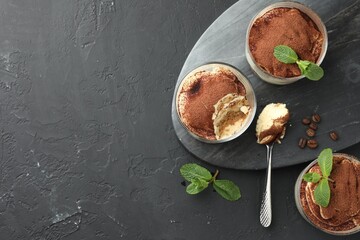 Delicious tiramisu in glasses, spoon, mint leaves and coffee beans on black table, top view. Space...