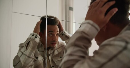 A young Black-skinned brunette man with stubble in a cream-colored checkered shirt preens near a mirror in the hallway in a modern apartment with light walls and good lighting
