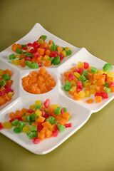 On yellow background, close-up dessert of colored candied fruit is laid out on white square-shaped plate. Sweet dessert in form candied fruit cubes close-up in a white menager on a yellow background
