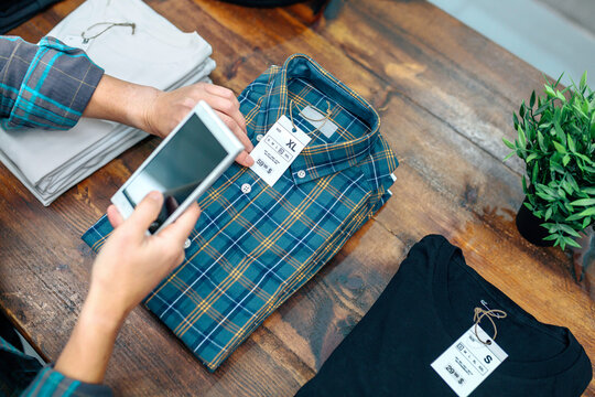 Unrecognizable woman customer holding cellphone while scan label with price and size of plaid shirt in clothes shop. Female employee taking photo of apparel stacks over counter on local store.