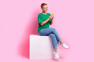 Full size photo of pleasant girl dressed green t-shirt jeans sit on white cube look at smartphone...